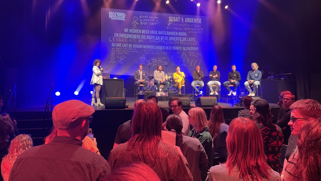 Paneldiscussie op een podium met zes panelleden zittend op krukken en een spreker. Tekst op achtergrond en publiek in de zaal.