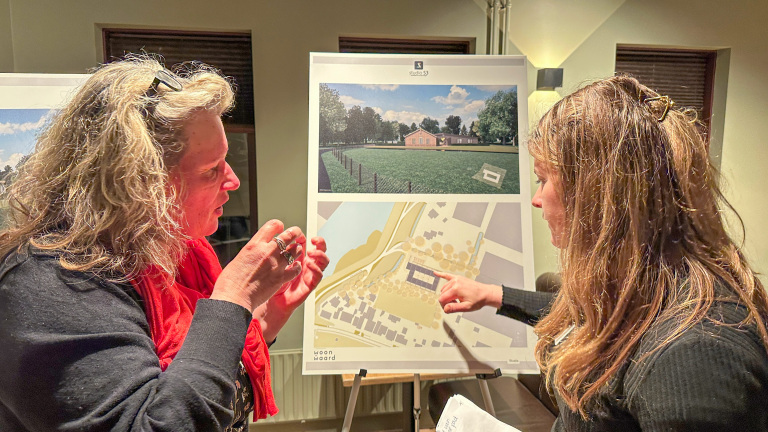 Twee vrouwen bespreken een poster met daarop een architecturale plattegrond en een buitenfoto.