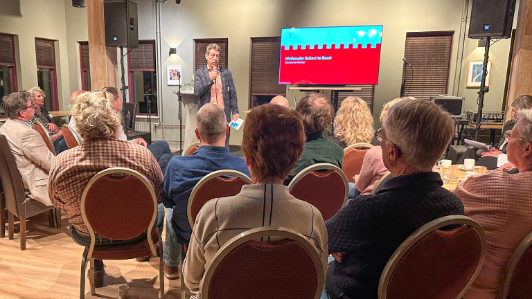 Man spreekt voor publiek met presentatie op scherm in zaal.