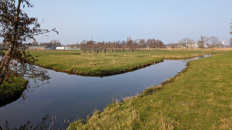 Een rustig landschap met een kronkelende beek, omgeven door groene weiden en kale bomen onder een heldere hemel.