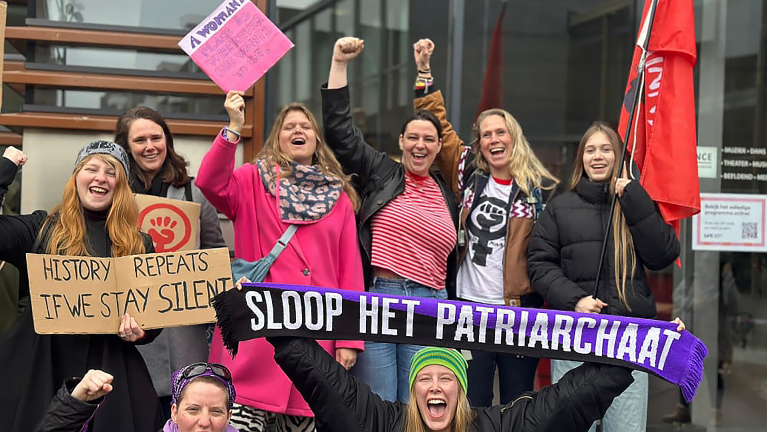 Groep mensen poseert enthousiast met protestborden en een sjaal met "Sloop het patriarchaat."
