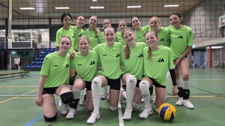 Een volleybalteam van jonge vrouwen poseert in een sporthal, in groene shirts met zwarte korte broeken.