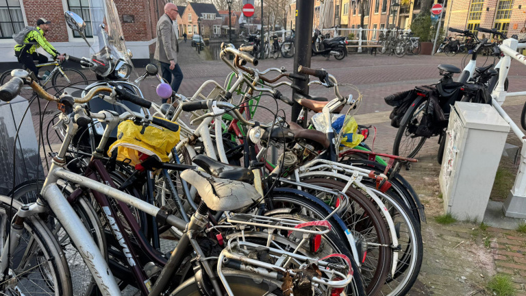 Een groep oude fietsen, waaronder een met een gescheurde zadel, geparkeerd op een steenachtige straat; mensen en andere fietsen op de achtergrond.