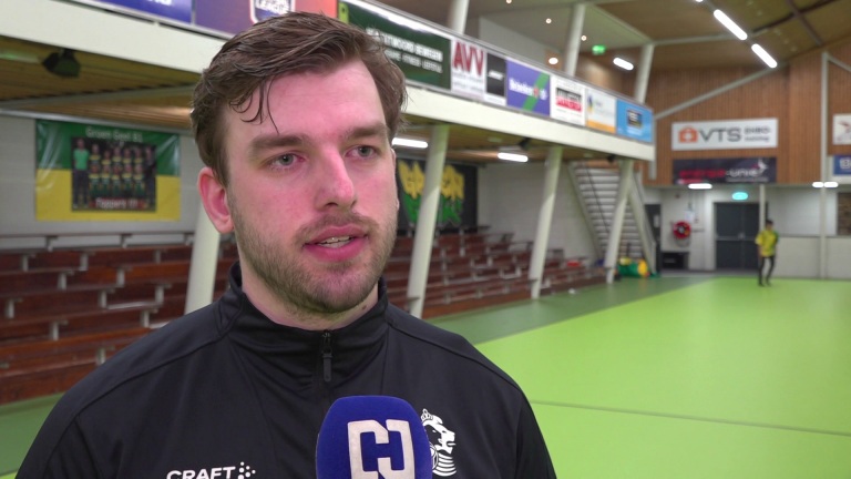 Een man in een sporthal geeft een interview; hij draagt een zwart sportjack en er is een microfoon met logo zichtbaar.