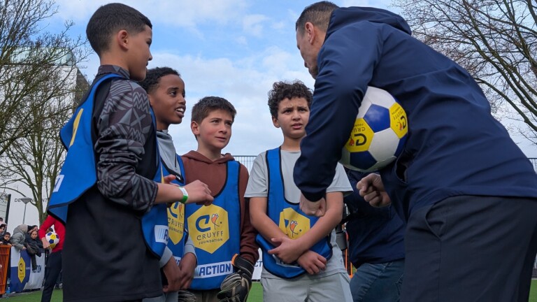 Een groep jongens in blauwe hesjes krijgt instructies van een man met een voetbal op een sportveld.