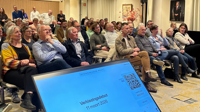 Mensen luisteren naar een verkiezingsdebat op 11 maart 2026; een scherm met details is op de voorgrond zichtbaar.