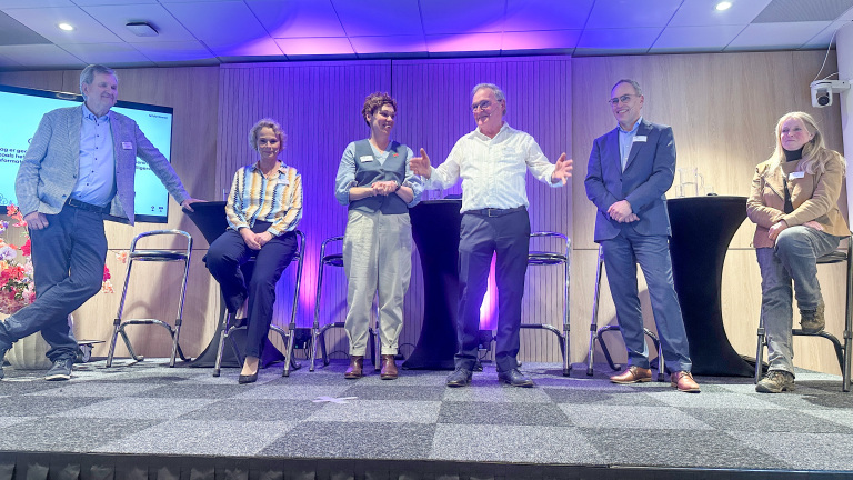 Zes personen staan op een podium in een zaal, waarvan drie staan en drie op krukken zitten, met een scherm op de achtergrond.