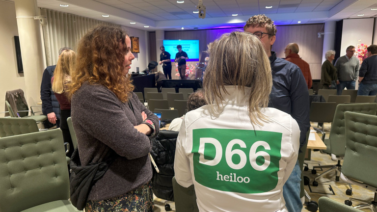 Mensen in gesprek in een zaal na afloop van een evenement; een persoon draagt een shirt met "D66 Heiloo".