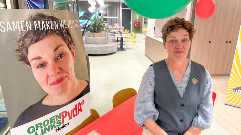 Vrouw zit naast GroenLinks PvdA-poster met haar portret en slogan "SAMEN MAKEN WE HEILOO" in een moderne ruimte.