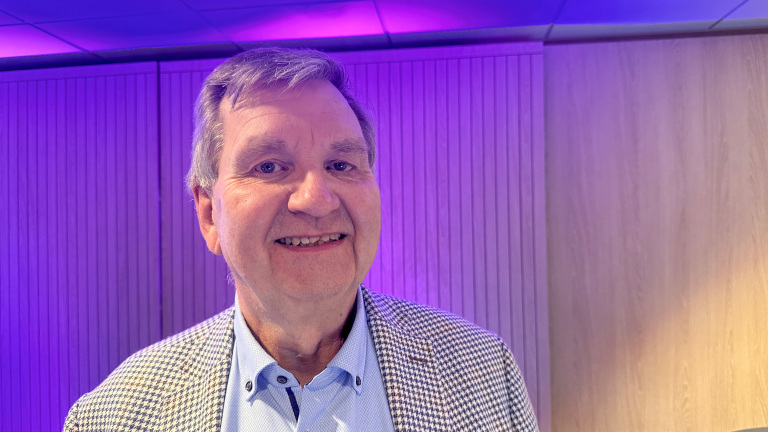 Man in een geruit jasje glimlacht voor een houten wand, met paars verlichte plafondpanelen op de achtergrond.