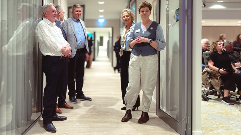 Mensen staan en zitten in een gang, enkele leunend tegen de muur, tijdens een bijeenkomst of evenement.