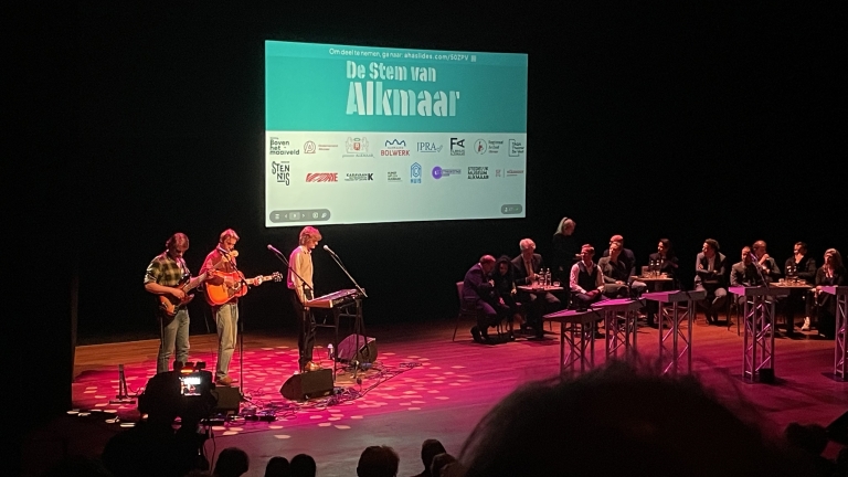 Muzikanten spelen op een podium met een projectiescherm erachter waarop "De Stem van Alkmaar" te zien is en een publiek luistert.