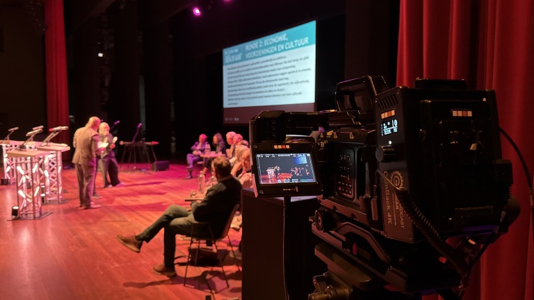 Een podium met mensen zittend op het toneel, gefilmd door een camera op de voorgrond.