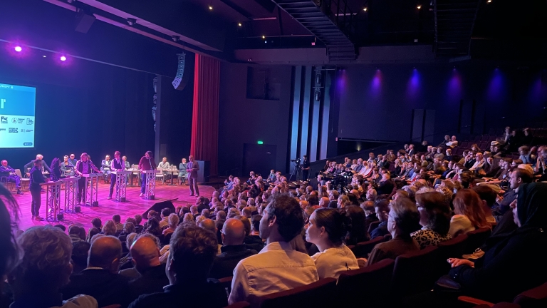 Een zaal vol mensen luistert naar een debat op een podium met meerdere sprekers achter lessenaars.