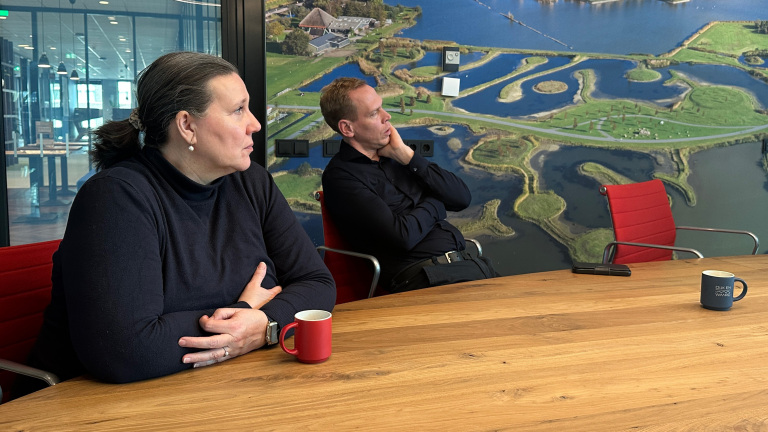 Twee personen zitten aan een houten tafel in een vergaderruimte met een luchtfoto op de muur.