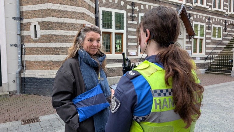 Een vrouw spreekt met een handhavingsmedewerker in uniform voor een historisch bakstenen gebouw.