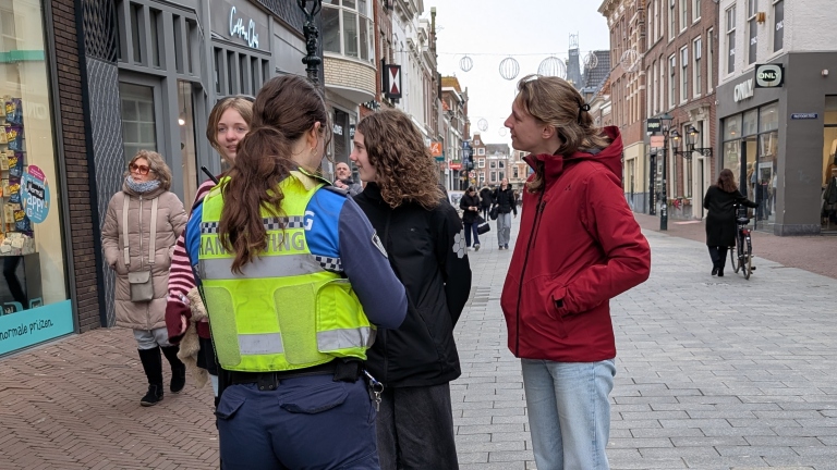 Een handhaver praat met twee personen op een winkelstraat, omringd door voorbijgangers.
