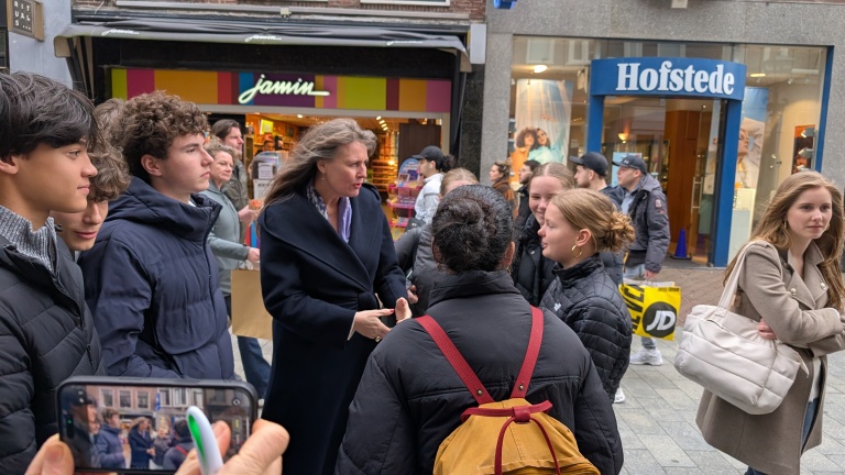 Mensen staan in een winkelstraat en praten met elkaar; op de achtergrond zijn winkels van Jamin en Hofstede te zien.