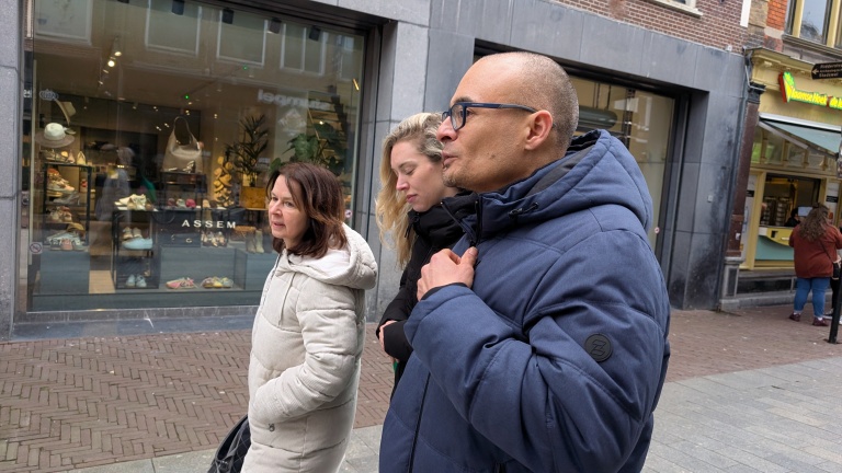 Drie mensen lopen voorbij een winkel met schoenen in de etalage, gekleed in warme jassen op een stenen trottoir.