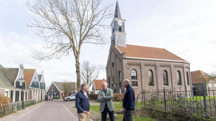 Drie mannen staan pratend voor een oude kerk met een torenspits in een dorpsomgeving.