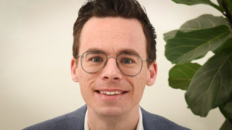 Man met bril en donker haar glimlacht naar de camera, met een plant op de achtergrond.