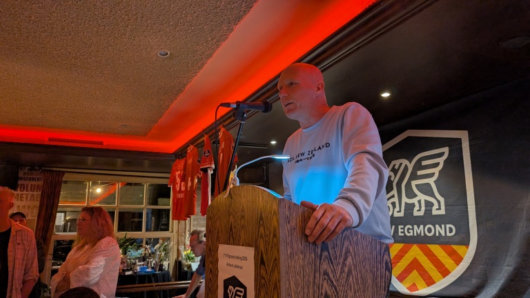 Man spreekt in microfoon op een podium in een ruimte met rode verlichting; shirts en een vlag zijn op de achtergrond zichtbaar.