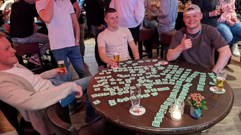 Drie mannen zitten aan een tafel vol met loten en bierglazen in een drukke bar; een van hen steekt zijn duim op.