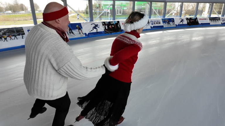 Twee mensen in warme kleding schaatsen samen binnen op een ijsbaan, waarbij de man de vrouw vasthoudt.