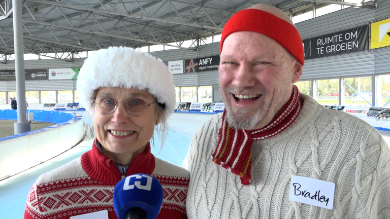 Twee lachende mensen in winterkleding bij een ijsbaan, met een microfoon voor hen.