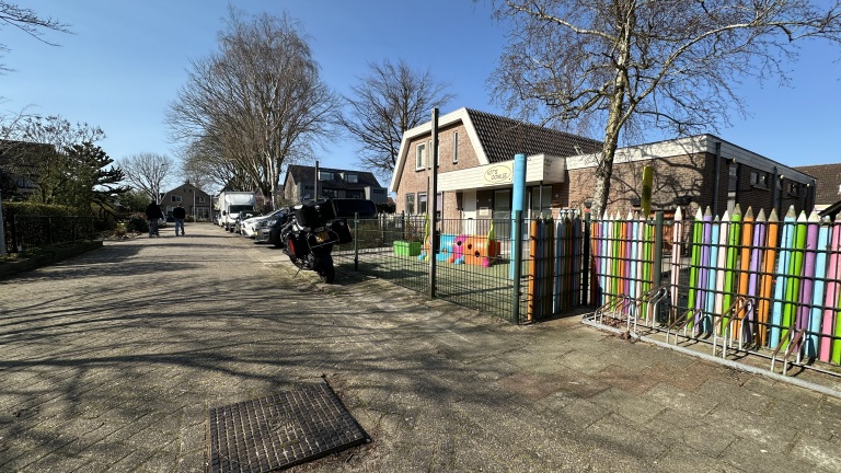 Kinderopvanggebouw met kleurrijk hek, speeltuinobjecten en enkele bomen op een zonnige dag. Straat met geparkeerde auto's.