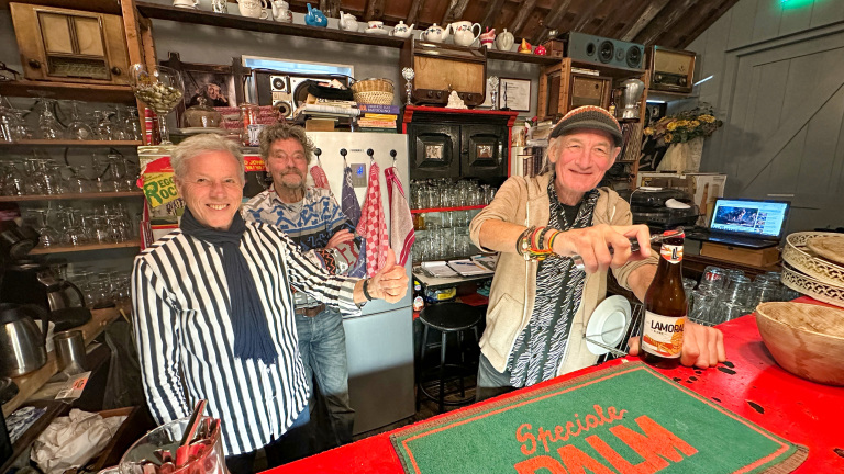 Drie lachende mannen staan achter een bar vol glazen en snuisterijen, met een man die een flesje bier opent.