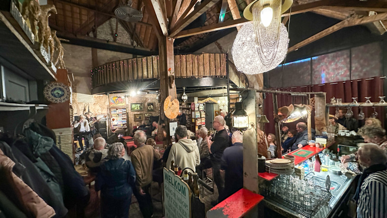 Mensen luisteren naar een muziekoptreden in een sfeervolle, rustieke bar met een houten interieur en gezellige verlichting.
