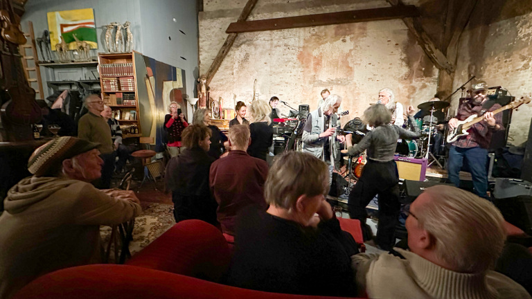 Een band speelt in een gezellige ruimte met publiek dat luistert en danst, omringd door boeken en decoratieve objecten.