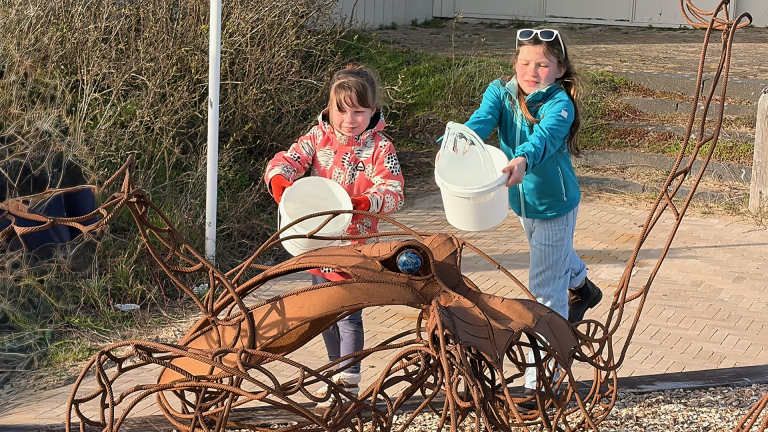 Twee kinderen dragen emmers bij een grote metalen sculptuur van een inktvis in een natuuromgeving.