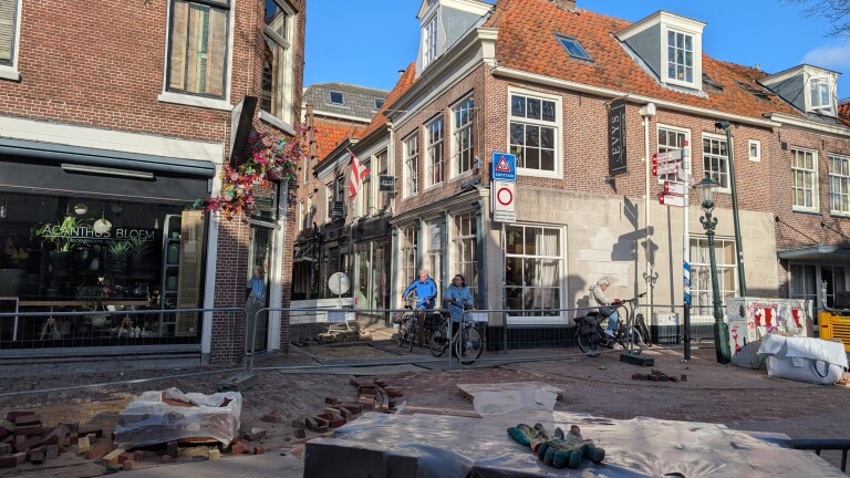 Straat met wegwerkzaamheden, bakstenen en omheining, naast een bloemenwinkel en fietsen geparkeerd tegen een historisch gebouw.