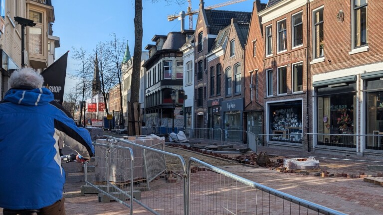 Straat in aanbouw met bouwhekken en stapels stenen, langs een rij historische gebouwen met winkels, op een zonnige dag.