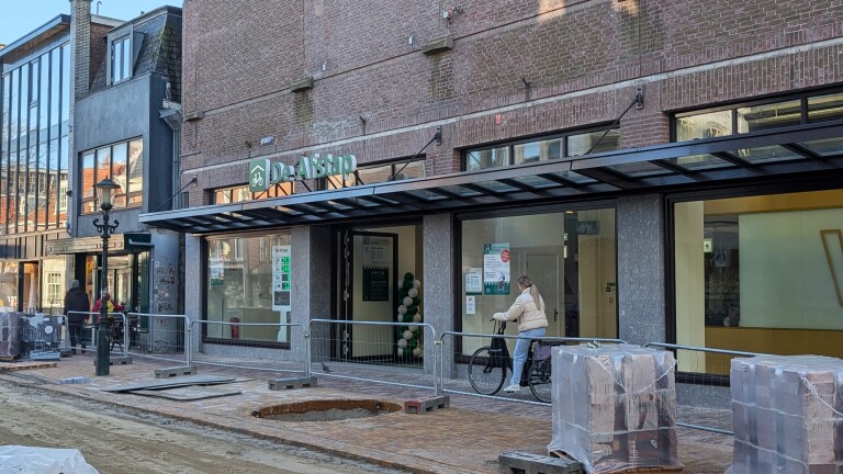 Een vrouw met een fiets voor een modern gebouw met een bordje van "De Aftrap" en straatwerken op de voorgrond.