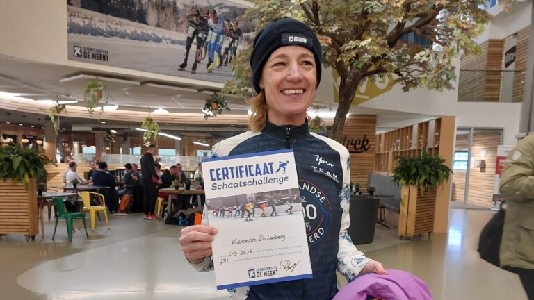 Vrouw in sportkleding houdt trots een certificaat vast in een sporthal met mensen op de achtergrond.