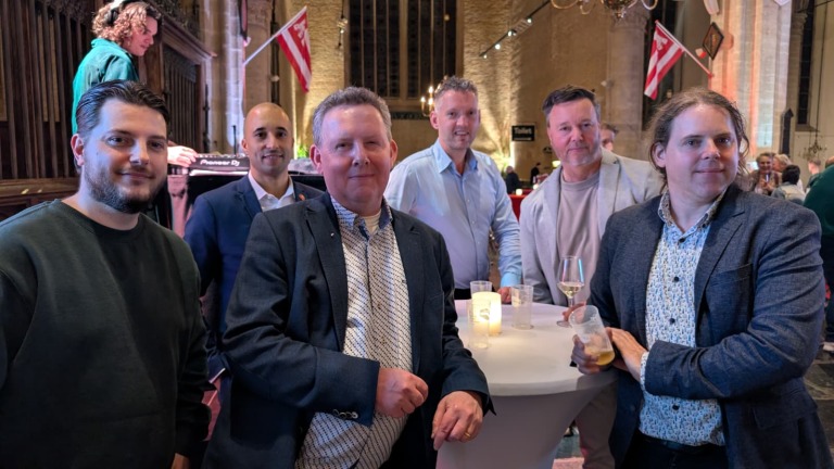 Zes mannen poseren bij een statafel in een feestelijke ruimte met gedimde verlichting en vlaggen, terwijl een DJ bezig is op de achtergrond.