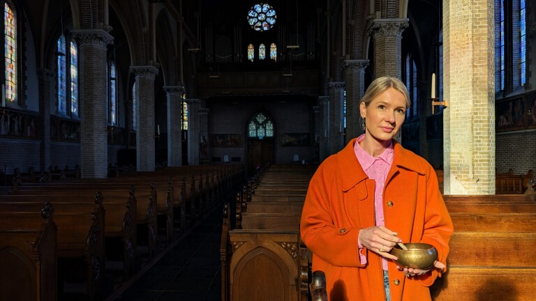 Vrouw in een oranje jas in een kerk, houdend een metalen kom met een klopper, omringd door houten banken en glas-in-loodramen.