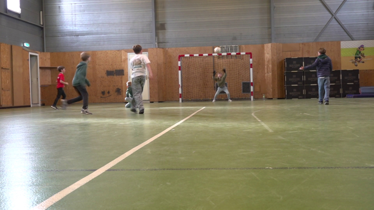 Kinderen spelen een potje zaalvoetbal in een sporthal, met één van hen die op doel schiet.