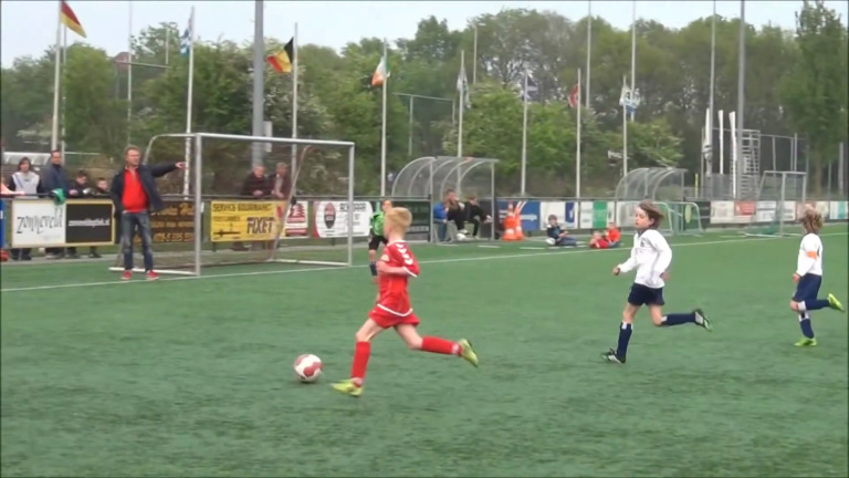 Kinderen spelen een voetbalwedstrijd op een kunstgrasveld; een speler in rood dribbelt de bal.