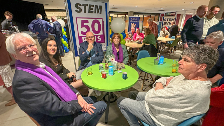 Groep mensen zit rond een felgroene tafel bij een campagne-evenement van 50PLUS, omringd door opvallende paarse sjaals.