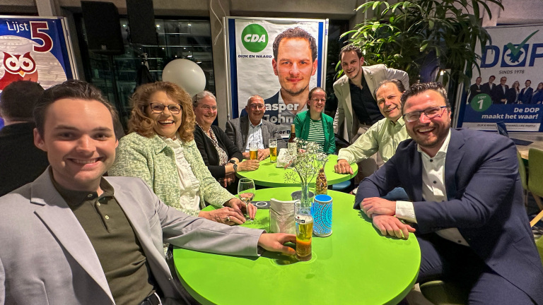 Groep mensen zit lachend aan groene tafels met drankjes, naast politieke campagneposters.