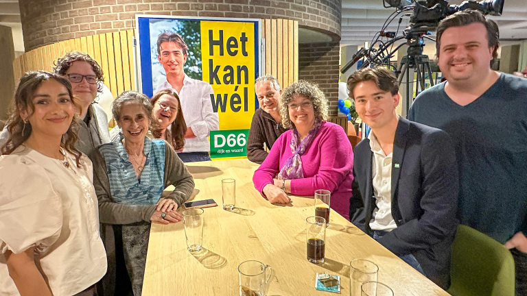 Een groep mensen lacht rond een tafel met glazen, naast een D66-poster met de tekst "Het kan wél".