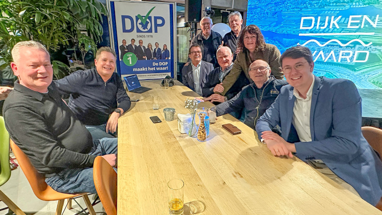 Groep mensen rond een tafel met een poster van de DOP-partij op de achtergrond, in een informele setting.