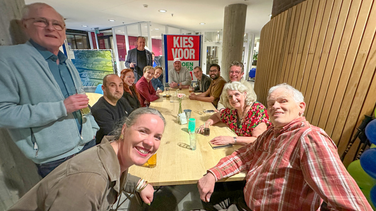 Een groep mensen zit aan een lange tafel in een binnenruimte, met een poster waarop "KIES VOOR" staat op de achtergrond.