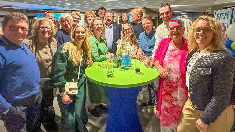 Groep mensen poseert vrolijk rond een hoge groene tafel, waarop drankjes en decoratie staan.
