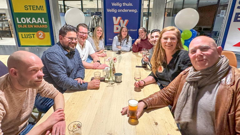 Een groep mensen zit lachend aan een tafel met drankjes, omgeven door verkiezingsborden.