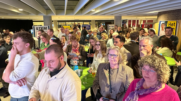 Een groep mensen verzameld in een zaal, waarvan sommigen gefocust zijn op hun telefoons.